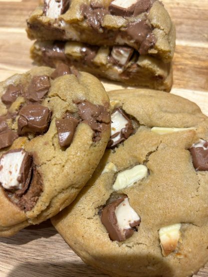 Three cookies with chunks of chocolate and marshmallow, stacked on a wooden surface.